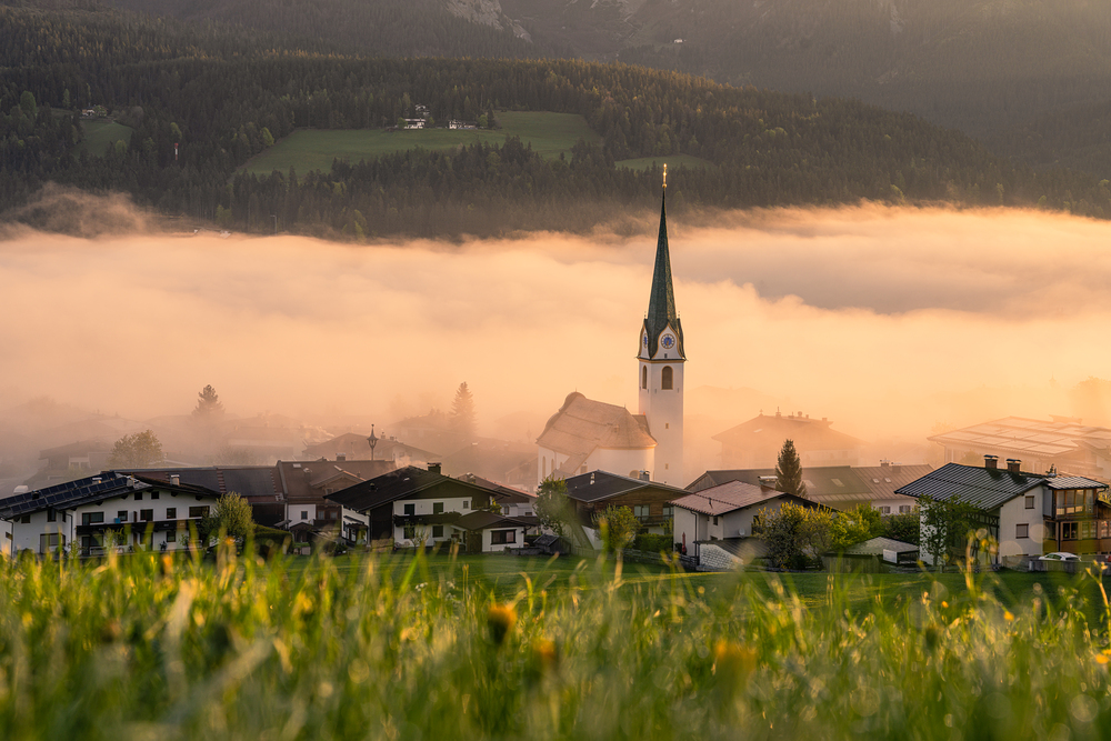 Blick auf das Dorf Ellmau in den frühen Morgenstunden. Über den Häusern liegt dichter Nebel, aus dem der hohe Kirchturm der Pfarrkirche deutlich herausragt. Die Szene ist von warmem Sonnenlicht in Gold- und Orangetönen geprägt. Im Vordergrund befinde